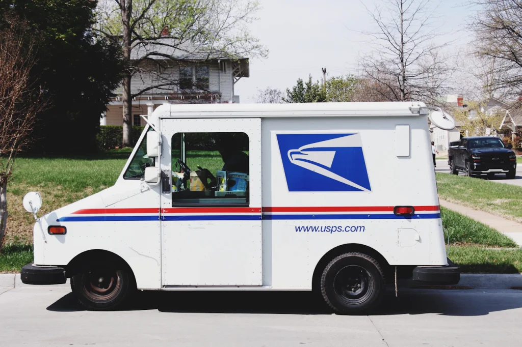 A US Postal Service mail carrier holding an Amazon package next to a white USPS delivery truck in a residential neighborhood.